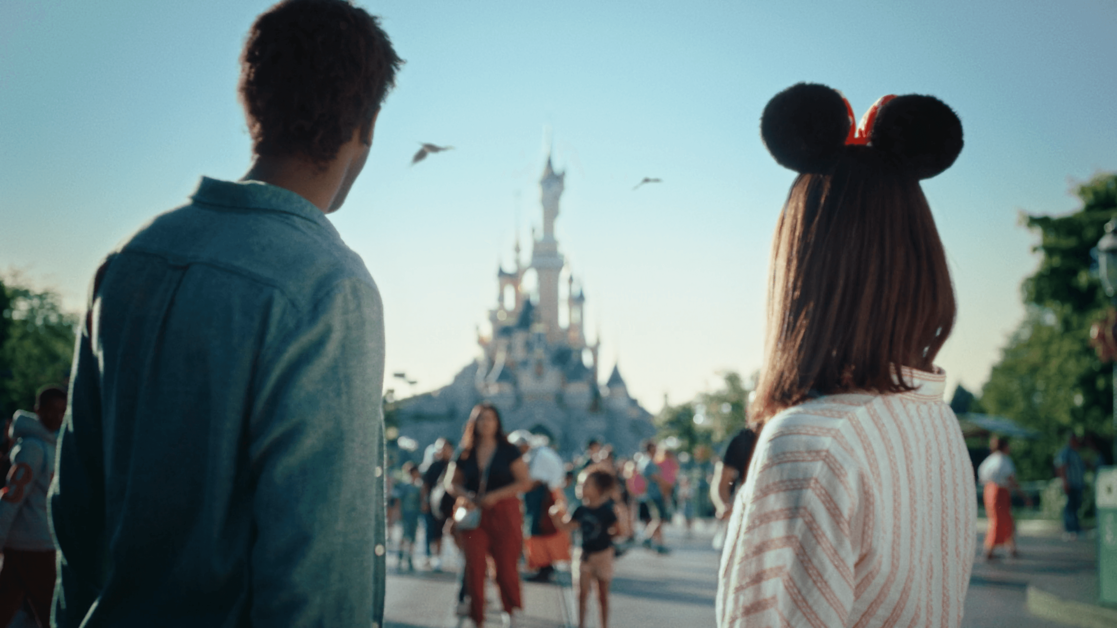 Two friends walking toward Sleeping Beauty Castle at Disneyland Paris at golden hour, one wearing Minnie Mouse ears