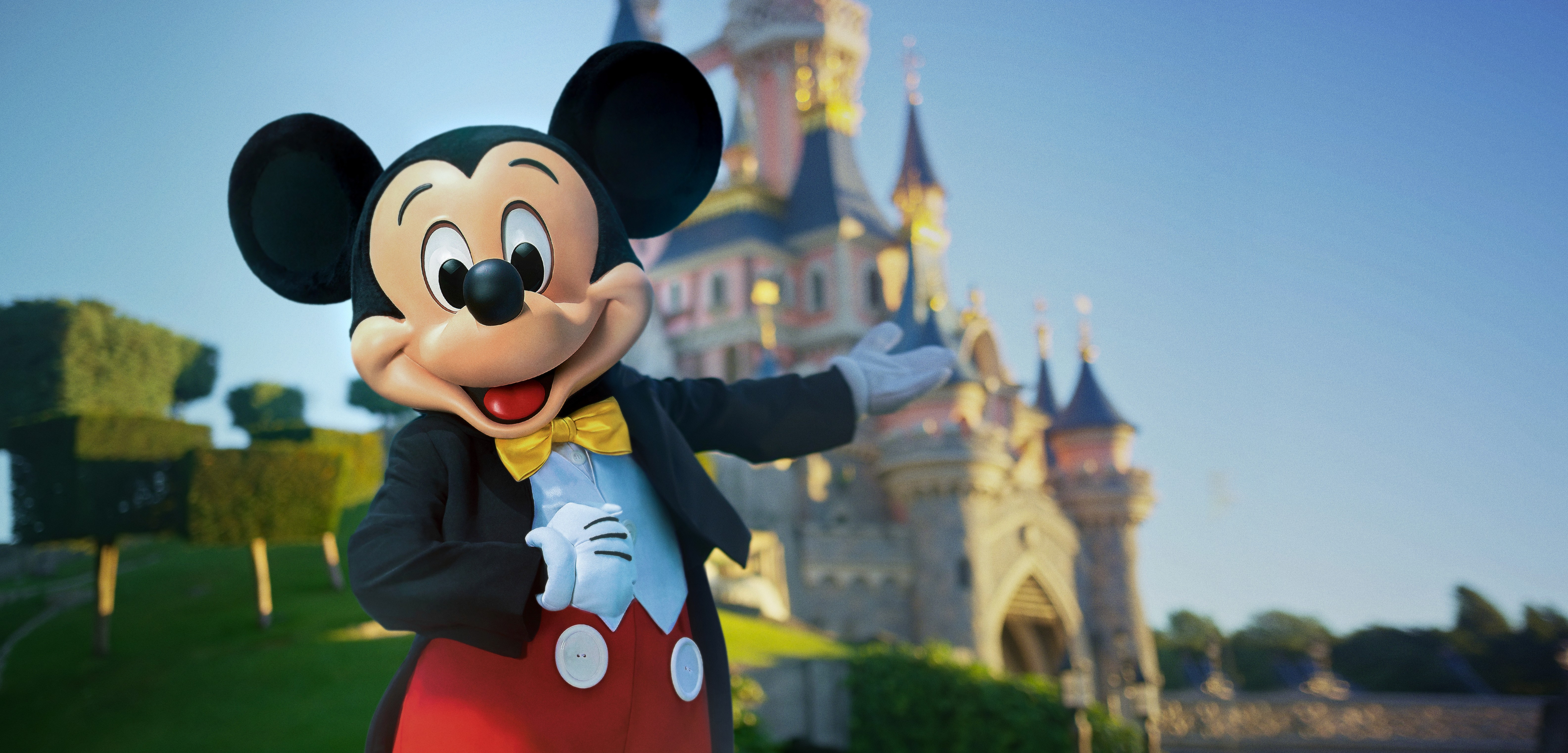 Mickey Mouse in a tuxedo gesturing toward Sleeping Beauty Castle at Disneyland Paris